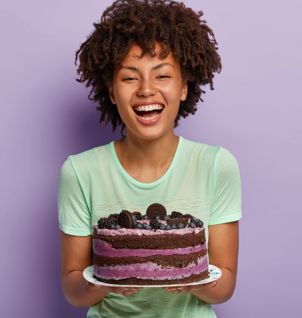 Happy birthday girl laughs joyfully, holds big tasty fruit cake, likes eating sweet food, improves mood with raising sugar in blood, has positive face look, stands over purple background, blank space